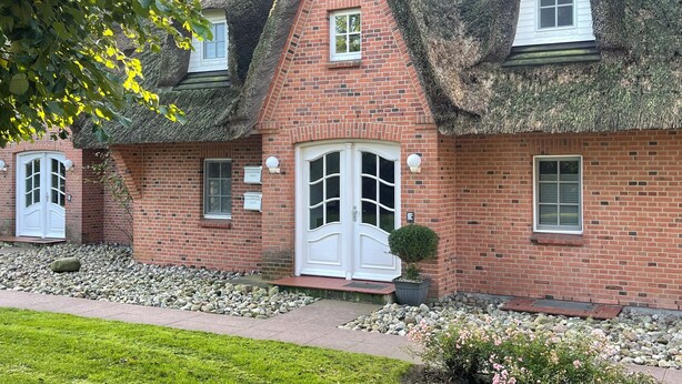 A traditional brick house with a thatched roof, a white front door, and a small garden with pebbles and flowers.