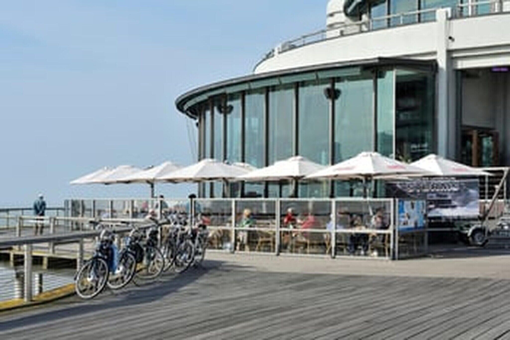 Een modern gebouw met een gebogen glazen gevel, buiten zitplaatsen met parasols, en fietsen die ervoor geparkeerd staan.