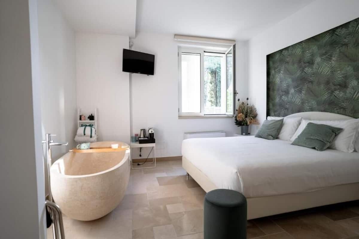 A modern bedroom with a large bed, a built-in bathtub, a television mounted on the wall, and a window with a view of greenery.