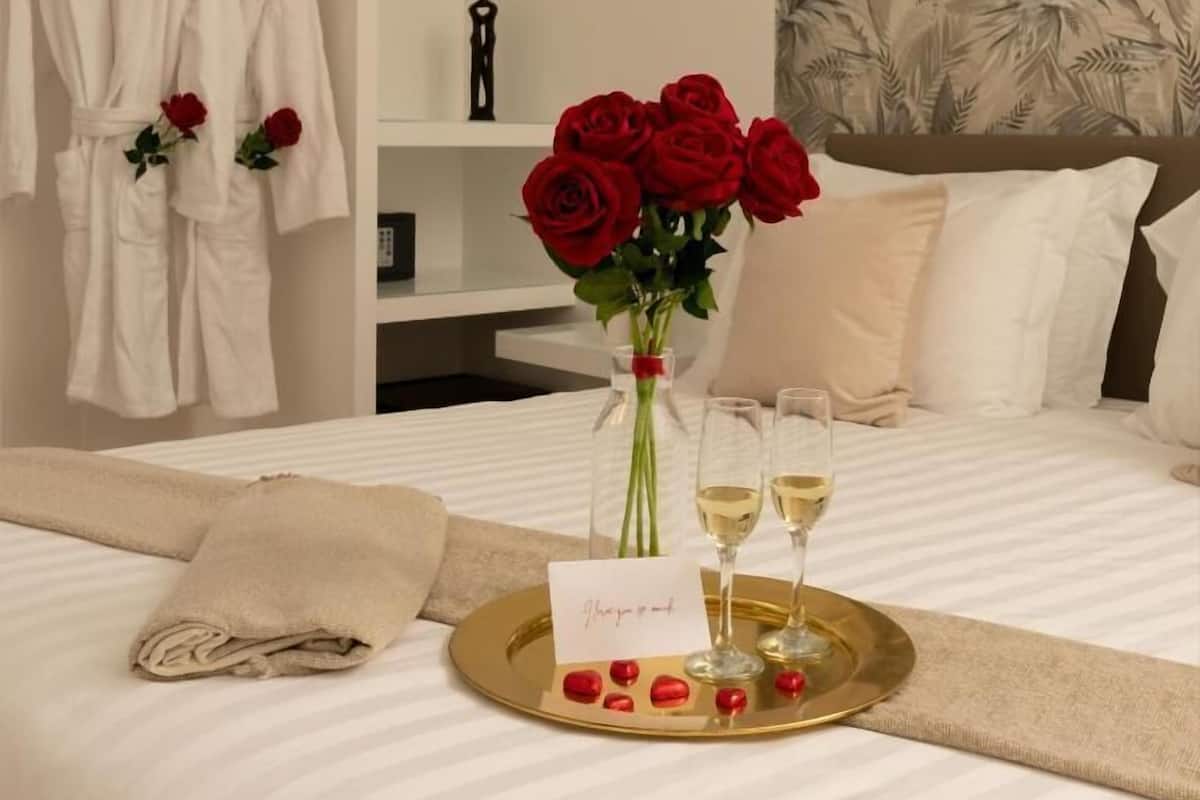 A hotel room with a bed, a robe rack, a vase with red roses, a tray with champagne glasses, and a card.