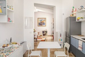 Una cocina moderna con muebles blancos, un comedor con mesa y sillas blancas, y un cuadro visible en la pared.