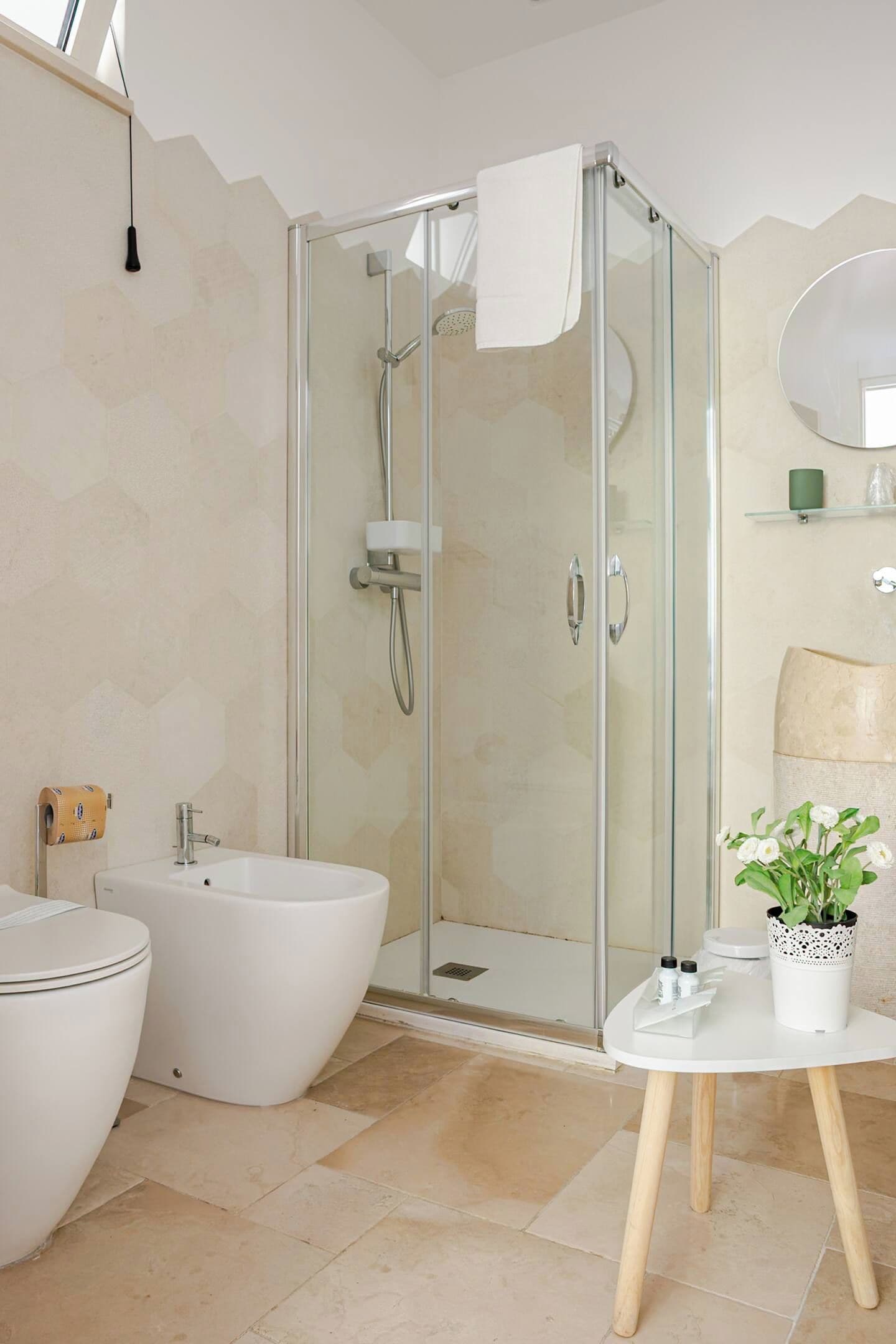 A modern bathroom with a glass-enclosed shower, a white toilet, a bidet, and a small table with a potted plant.