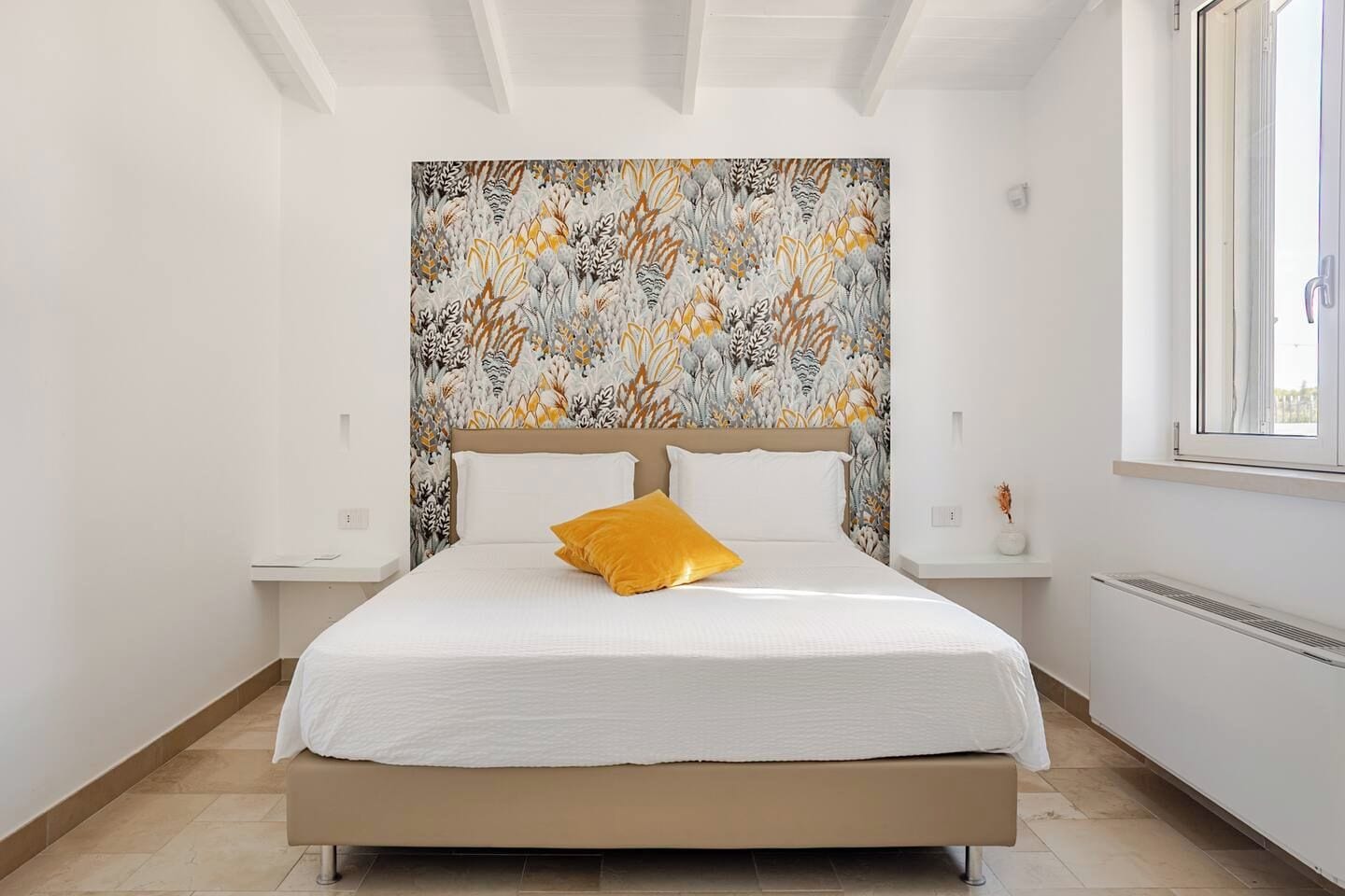 A bedroom with a large bed, a headboard with a floral pattern, a window, and a radiator.