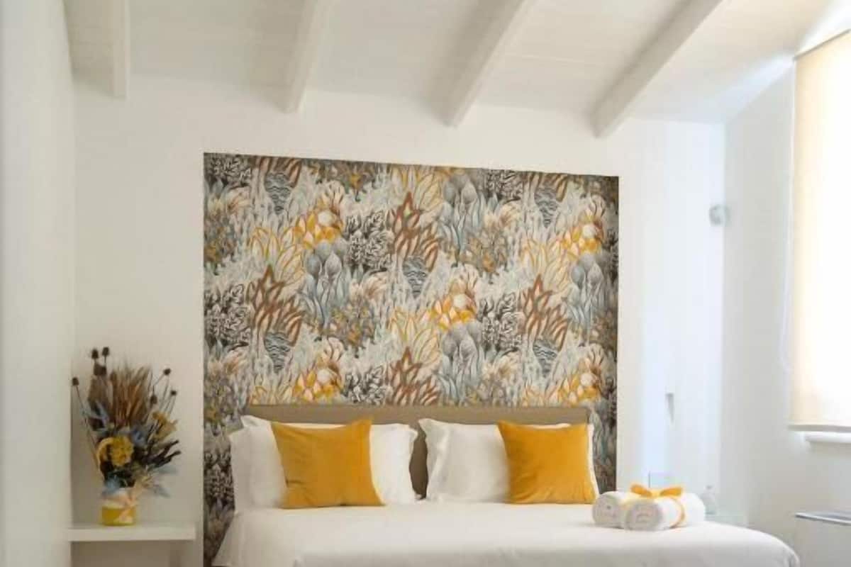 A bedroom with a bed, a chair, a wall with a floral pattern, and a window.