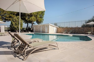 Piscina con sillones de descanso y sombrillas, rodeada por un muro de piedra y una valla metálica.