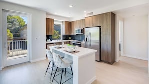 Une cuisine moderne dotée d’un îlot central, d’appareils électroménagers en acier inoxydable et d’une porte-fenêtre coulissante donnant sur l’extérieur.
