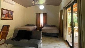 A hotel room with two beds, a sofa, a ceiling fan, a painting, and a large sliding glass door leading to an outdoor area.