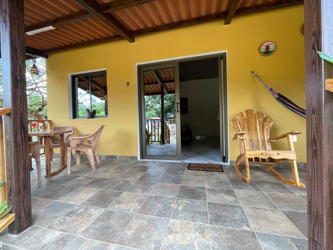 A covered patio area with tiled flooring, wooden furniture, and a wooden rocking chair.