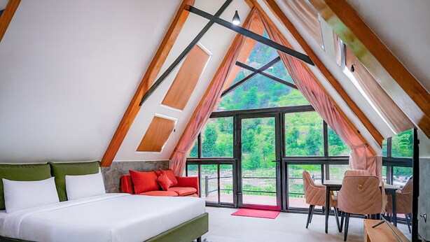 A bedroom with a bed, a sofa, a desk, and a chair, with large windows and a view of greenery. - Roameo Resorts (Sharda)