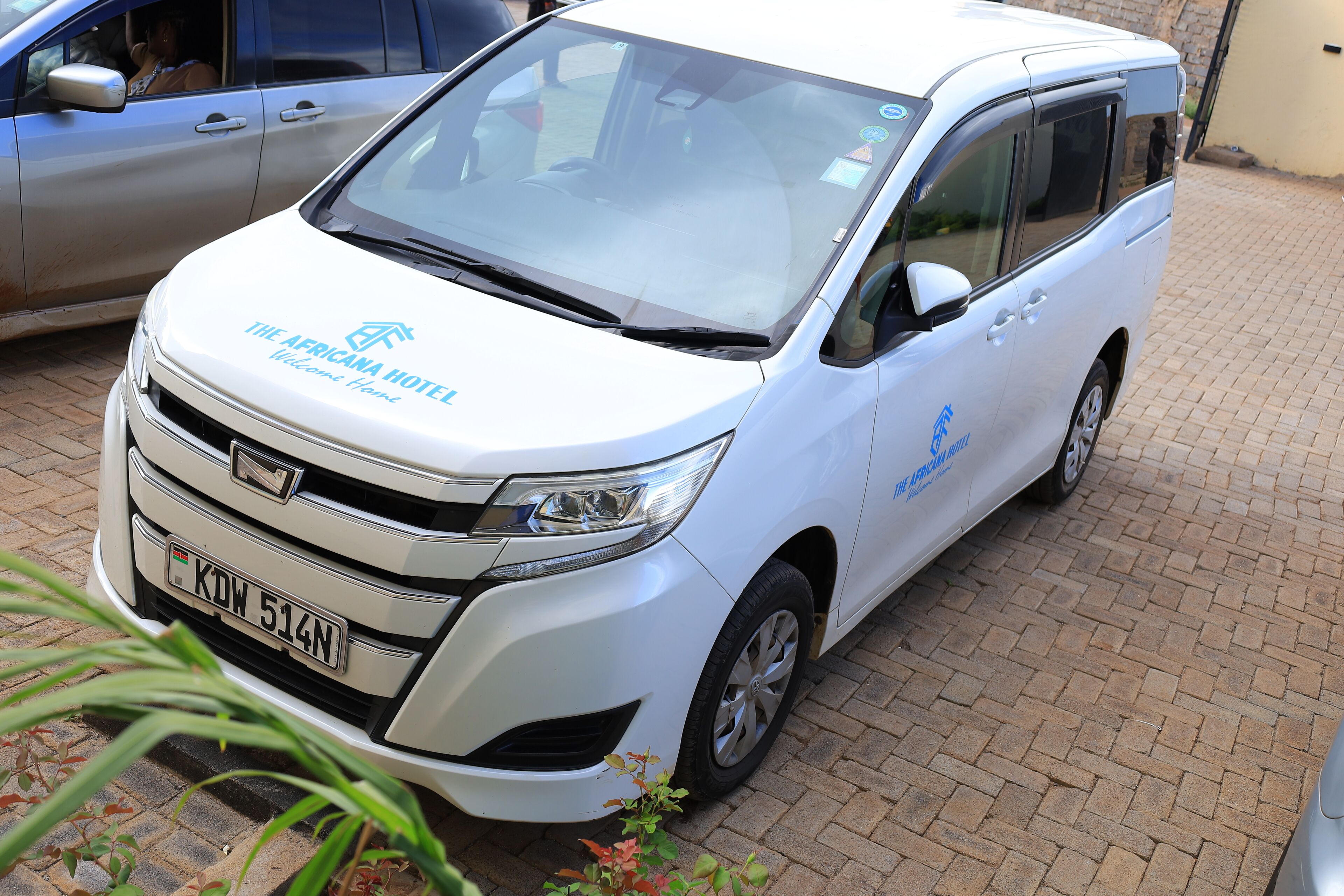 Uma minivan branca com a marca 'The Africana Hotel' estacionada em um piso de tijolos.