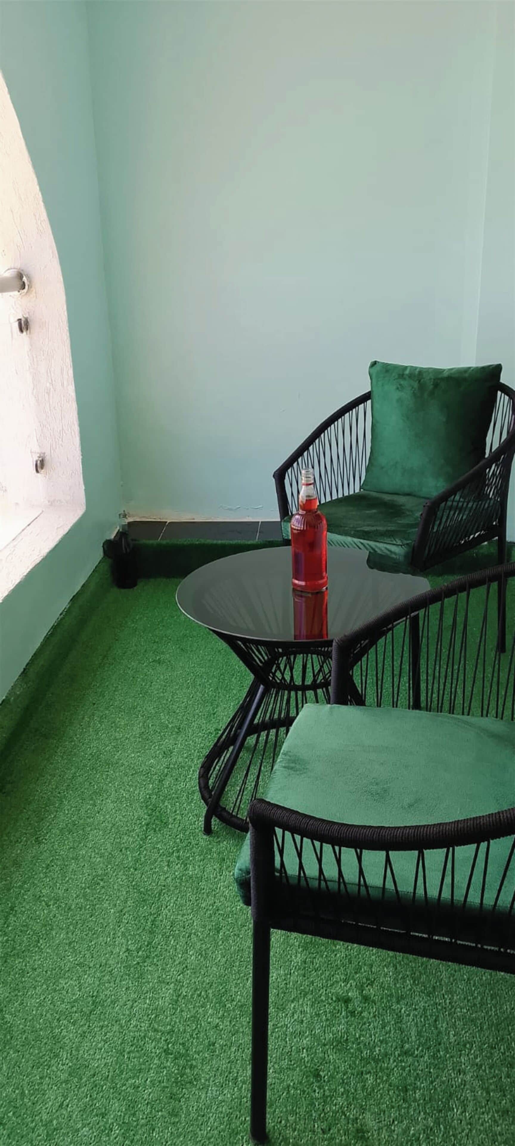 Una stanza con pavimento verde sintetico, un tavolo rotondo con una bottiglia rossa e mobili in vimini nero.