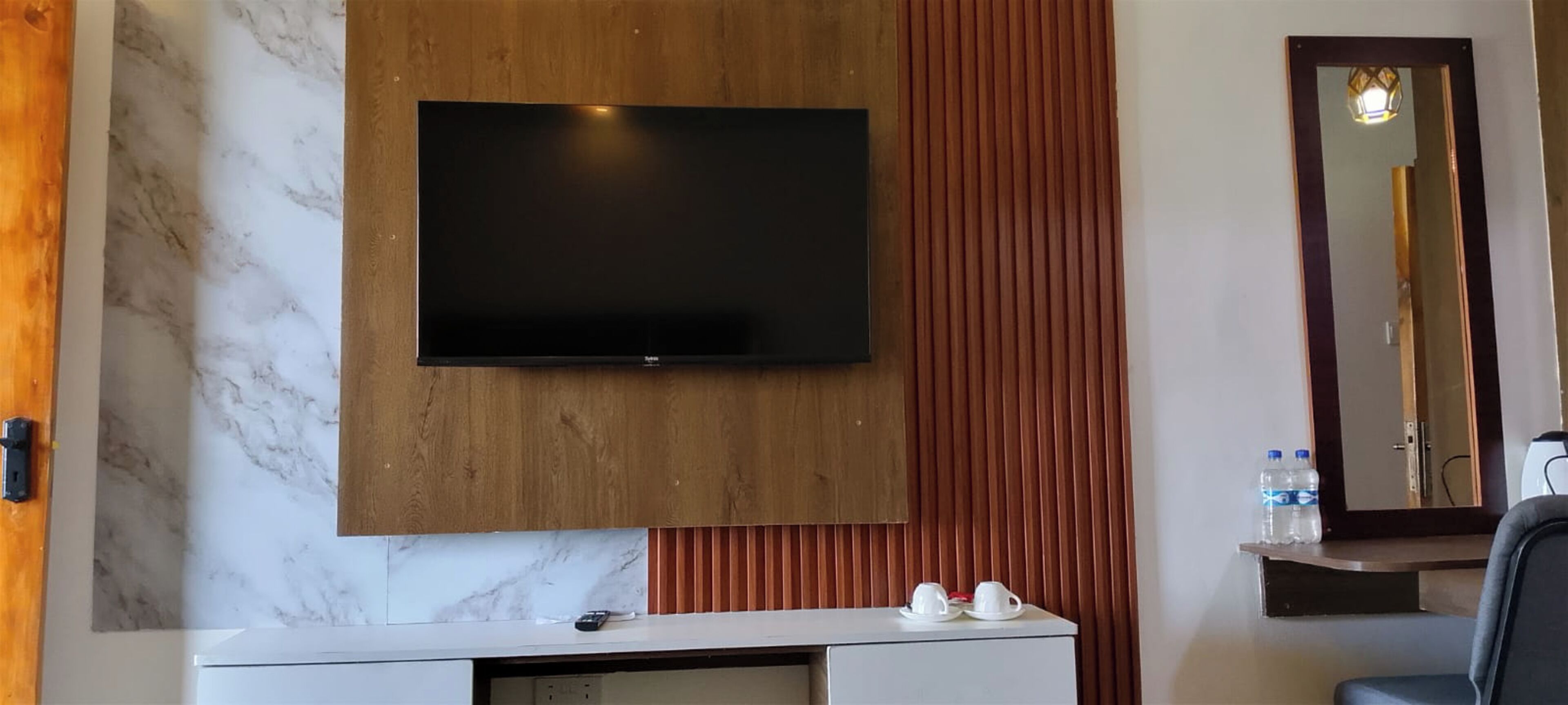 Hotelkamer met een flatscreen televisie bevestigd aan een houten paneel, een marmeren spatwand, een witte console tafel met een afstandsbediening, twee witte kopjes en een spiegel met houten lijst.