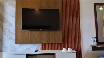 A hotel room with a flat-screen TV mounted on a wooden panel, a marble backsplash, a white console table with a remote control, two white cups, and a mirror with a wooden frame.