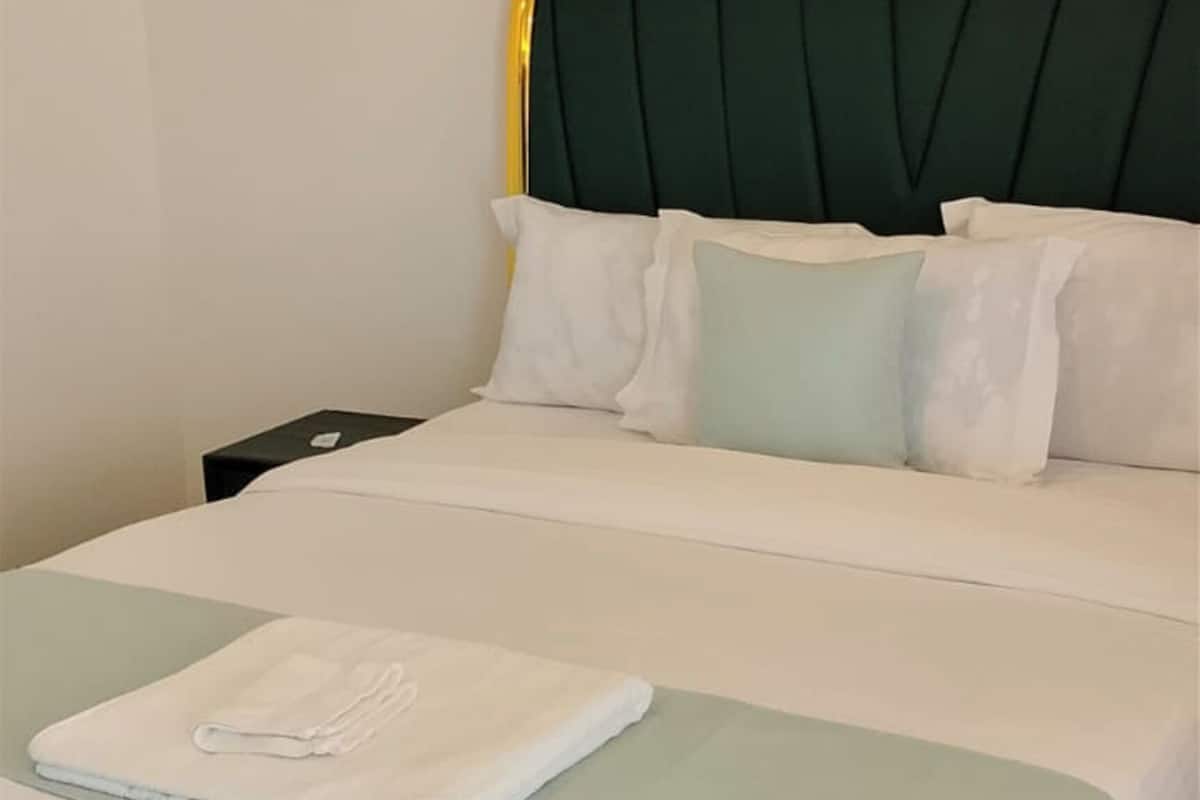 Ein ordentlich bezogenes Bett mit weißer Bettwäsche und einem grünen Kopfteil, ein gefaltetes weißes Handtuch auf dem Bett und ein kleiner Nachttisch mit einem schwarzen Gegenstand darauf.