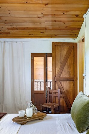 A wooden ceiling, a bed with a green pillow, a wooden door, a chair, and a tray with a pitcher and cups.