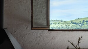 Une pièce avec une fenêtre donnant sur un paysage verdoyant, un tableau encadré au mur et un bureau avec des livres et une lampe.