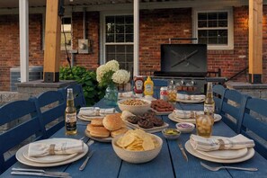 A dining table set outdoors with food, drinks, and a TV in the background.