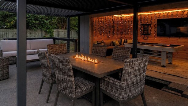 Une terrasse extérieure dotée d'un foyer, de meubles en osier, d'un canapé et d'un téléviseur fixé à un mur en bois.