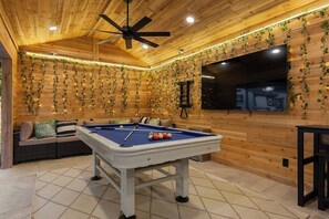 A wooden interior with a pool table, ceiling fan, and a sofa with cushions.