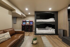 A room with a brown leather sofa, a coffee table with a plant, a bed with white bedding, and arcade games.