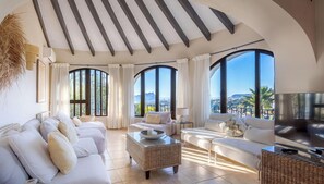 A spacious living room with a large sofa, a coffee table, and a TV. There are large windows offering a view of the outdoors. - Villa COSTA AZZURRA by Abahana Villas (Benissa)