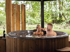 A wooden hot tub with a person and a bottle of wine on a table. - Experience luxury, tranquility, and wellness in the Nature Villa for 2 people with a private sauna, hot tub, and a unique nature experience. (Wekerom)