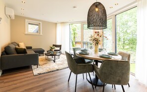 Ein modernes Wohnzimmer mit einer Couch, Sesseln, einem Esstisch und einem großen Fenster mit Blick auf Grünflächen.