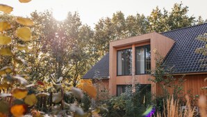 Una casa moderna de madera con techo inclinado, ventanas amplias y rodeada de árboles y vegetación.