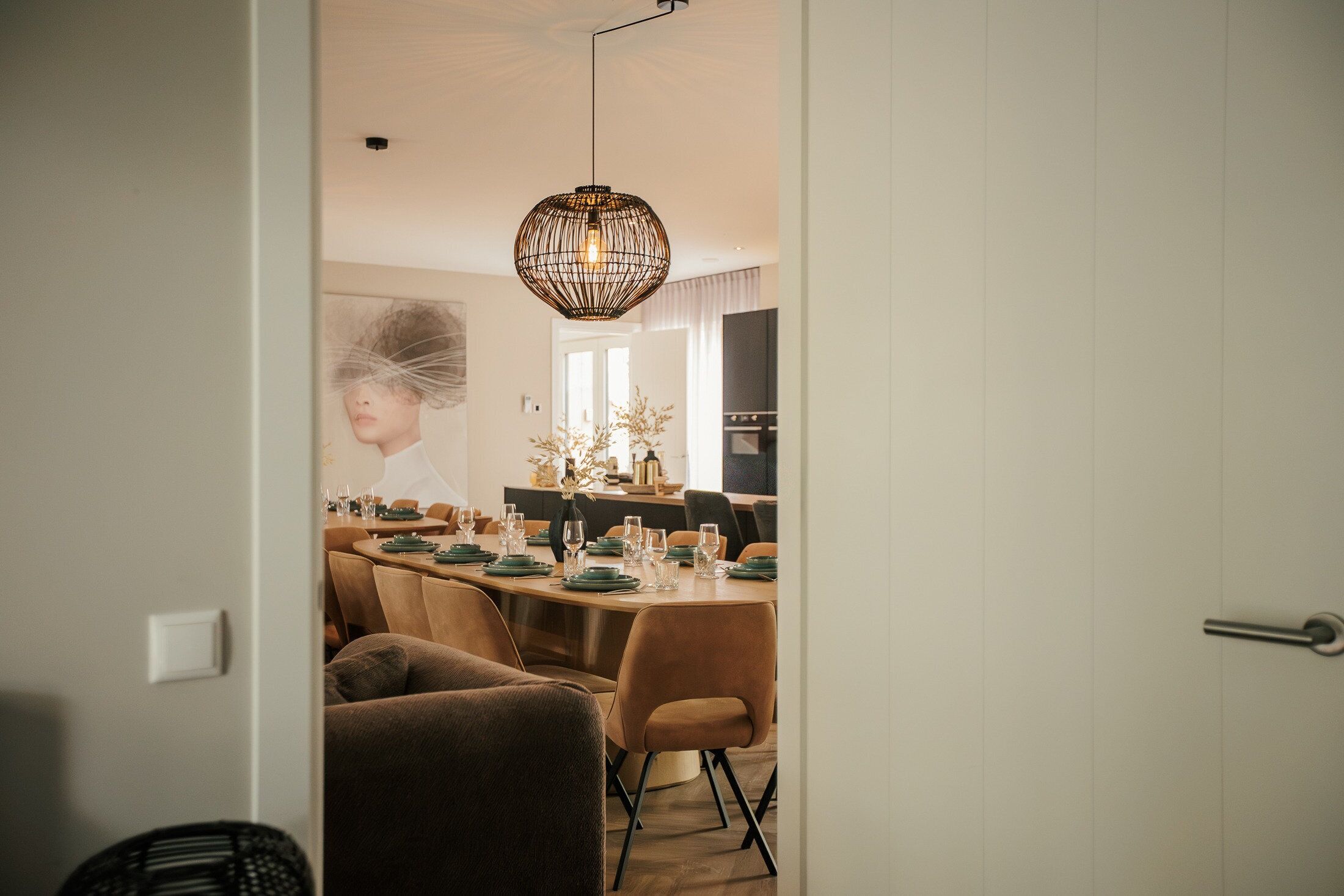 A modern dining area with a long table set for multiple people, a pendant light, and a view into a kitchen area.