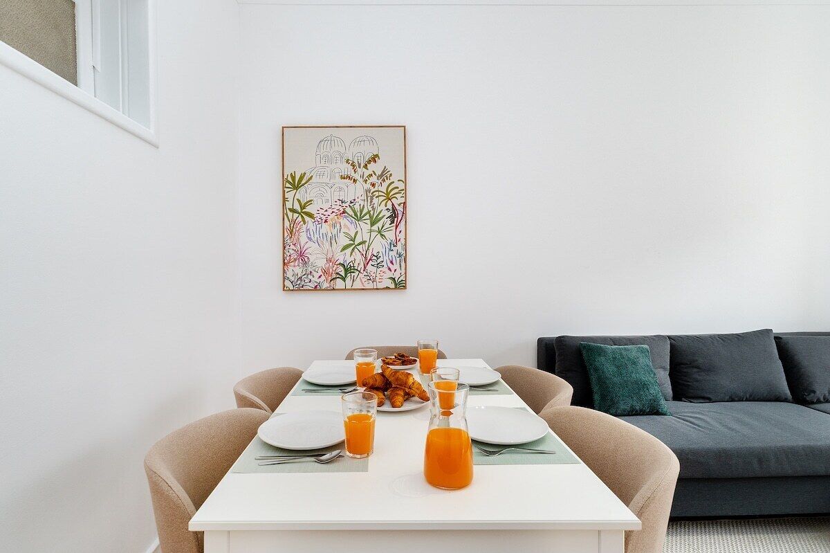 Et spiseområde med et bord dækket til to personer, et indrammet botanisk kunstværk på væggen og en mørk sofa i baggrunden.