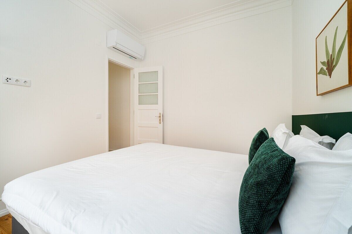 Een hotelkamer met een bed, een groen sierkussen, een witte muur, een ingelijste botanische print en een airconditioningsunit.