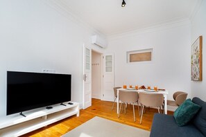 Ein modernes Wohnzimmer mit Flachbildfernseher, Essecke mit Tisch und Stühlen sowie einer Couch mit Kissen.