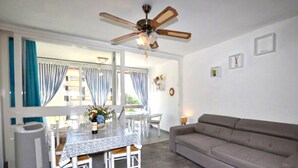 A living room with a grey sofa, a dining table with chairs, a ceiling fan, and a view through a large window with curtains. - Stunning apartment in Saint-Cyprien Plage (Saint-Cyprien Plage)