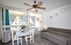 Ein Wohnzimmer mit grauem Sofa, einem Esstisch mit Stühlen, einem Deckenventilator und Blick durch ein großes Fenster mit Vorhängen.