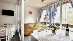 A room with a sofa, a dining table with a bottle of wine, a TV mounted on the wall, and a window with curtains. - Stunning apartment in Saint-Cyprien Plage (Saint-Cyprien Plage)