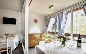 Ein Zimmer mit einer Couch, einem Esstisch mit einer Weinflasche, einem an der Wand montierten Fernseher und einem Fenster mit Vorhängen.