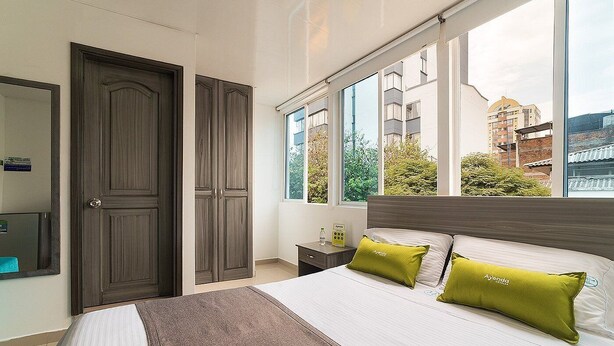 Ein Hotelzimmer mit einem großen Bett, einem Schrank, einem Nachttisch und einem Fenster mit Blick auf Gebäude und Bäume.
