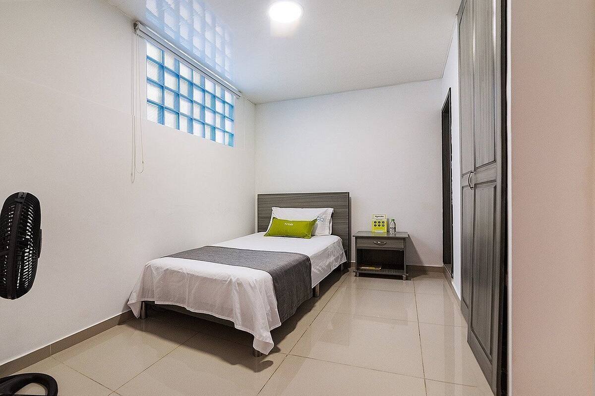 A small, clean room with a bed, a nightstand, a fan, and a window with blinds.