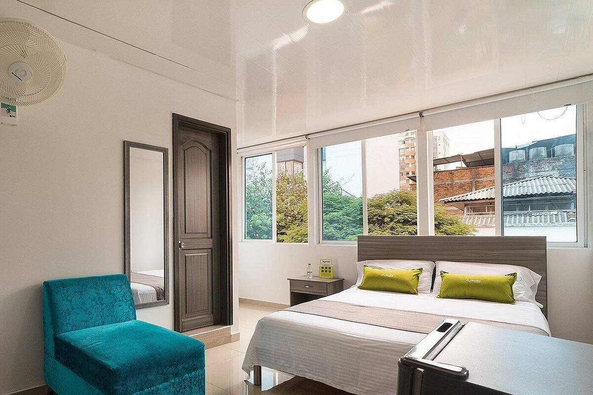 A bedroom with a bed, a chair, a nightstand, and a window with a view of buildings and trees.