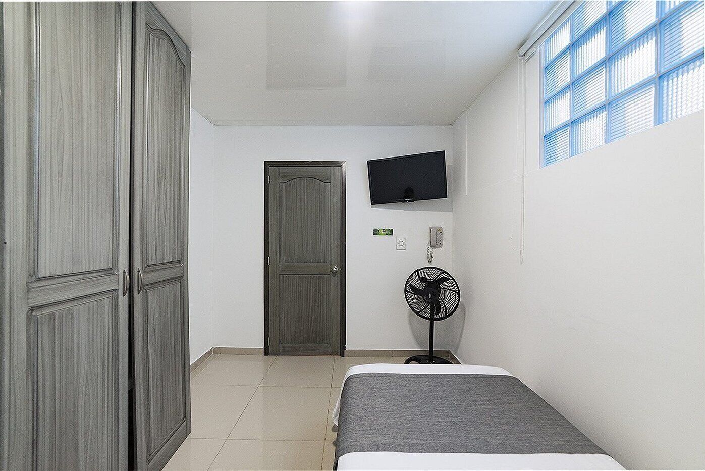 A small room with a bed, a wardrobe, a television, and a standing fan.