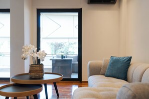 A modern living room with a sofa, a small round table with a vase of flowers, and a view through a glass door to an outdoor area. - Olive Glow, Glyfada Luxury Suites & Apartments (Athens)