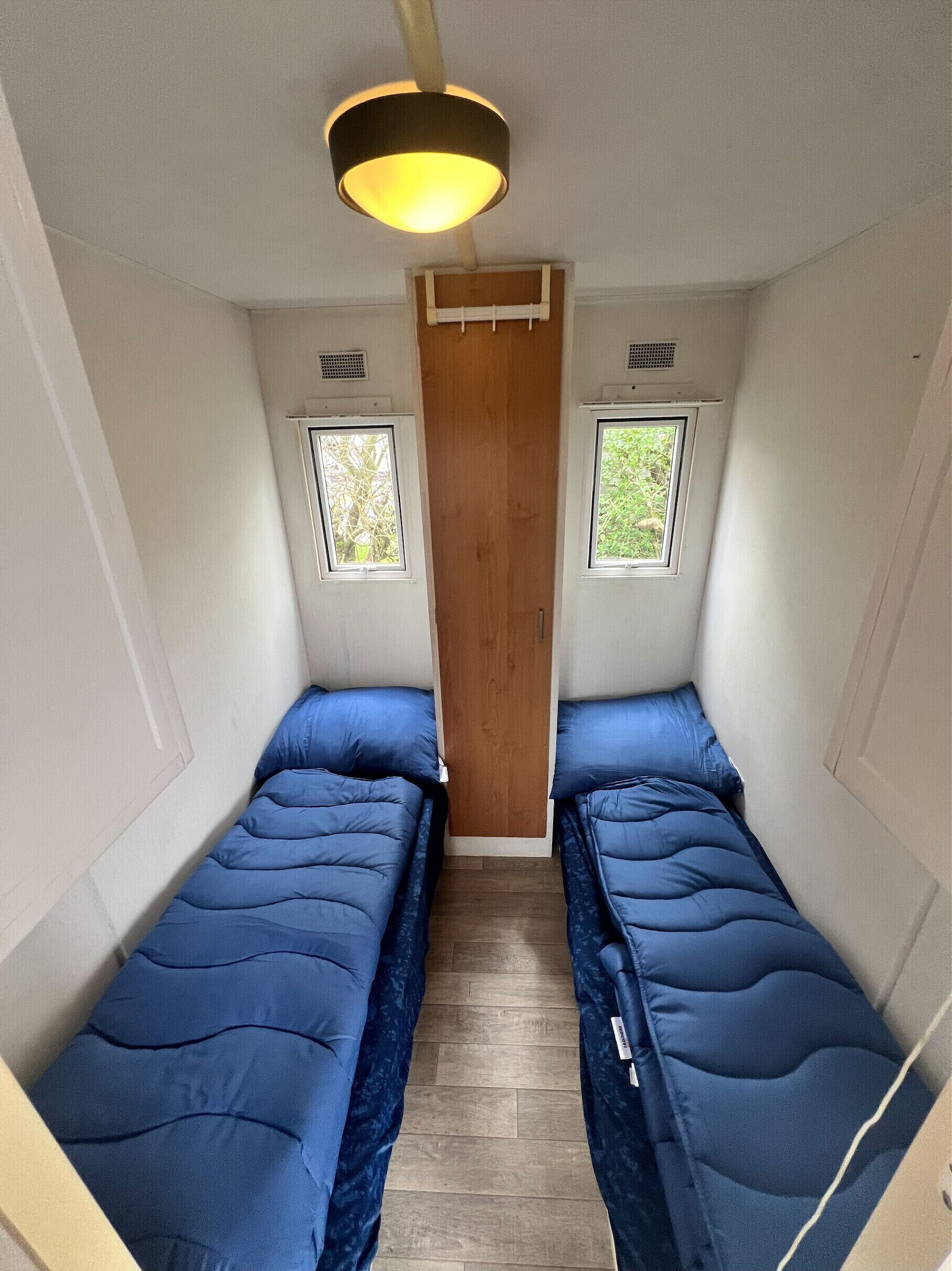 Habitación pequeña con dos camas individuales, una puerta de madera y dos ventanas pequeñas.