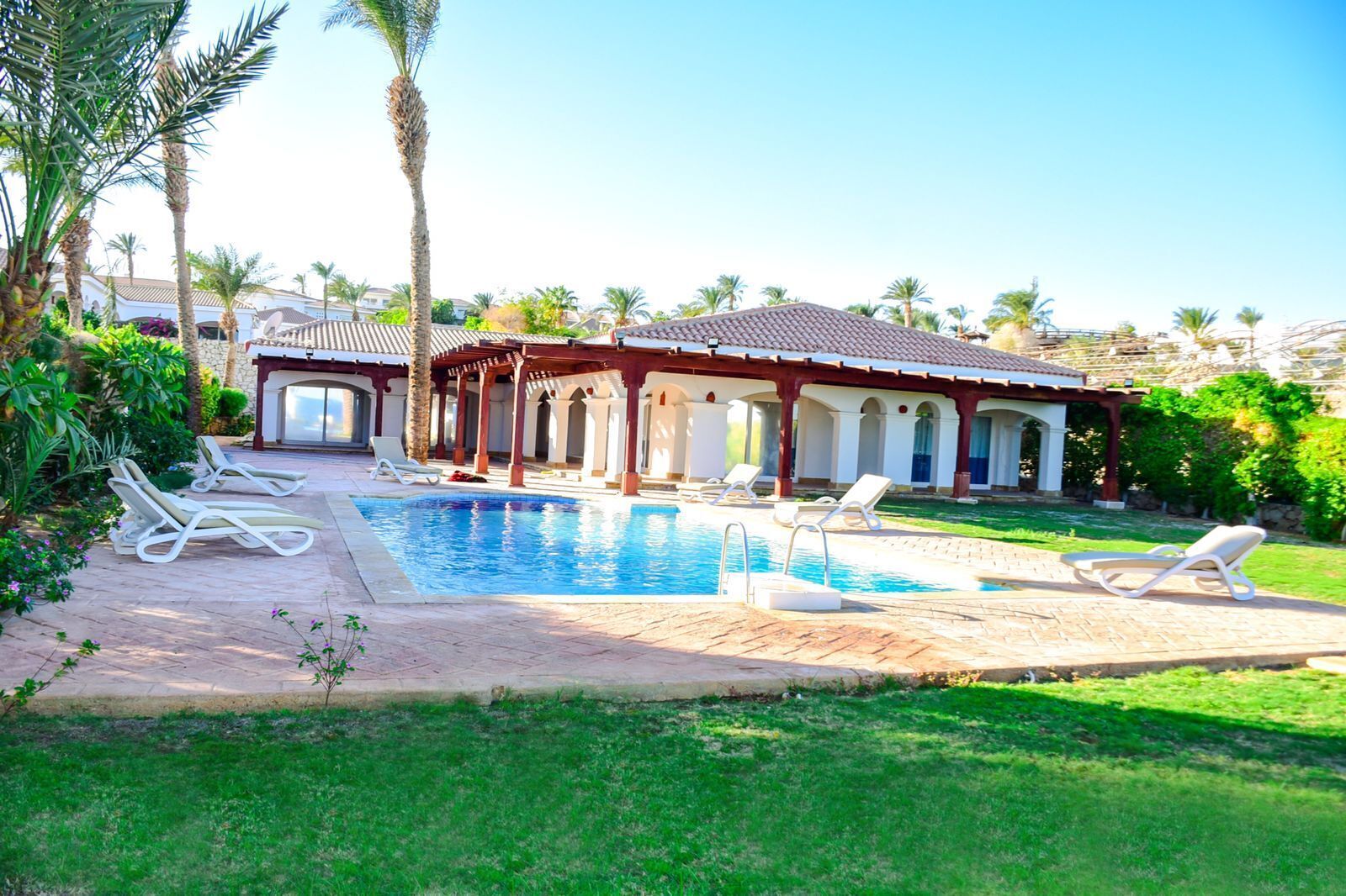 A pool area with lounge chairs, a covered patio, and a building with a tiled roof.