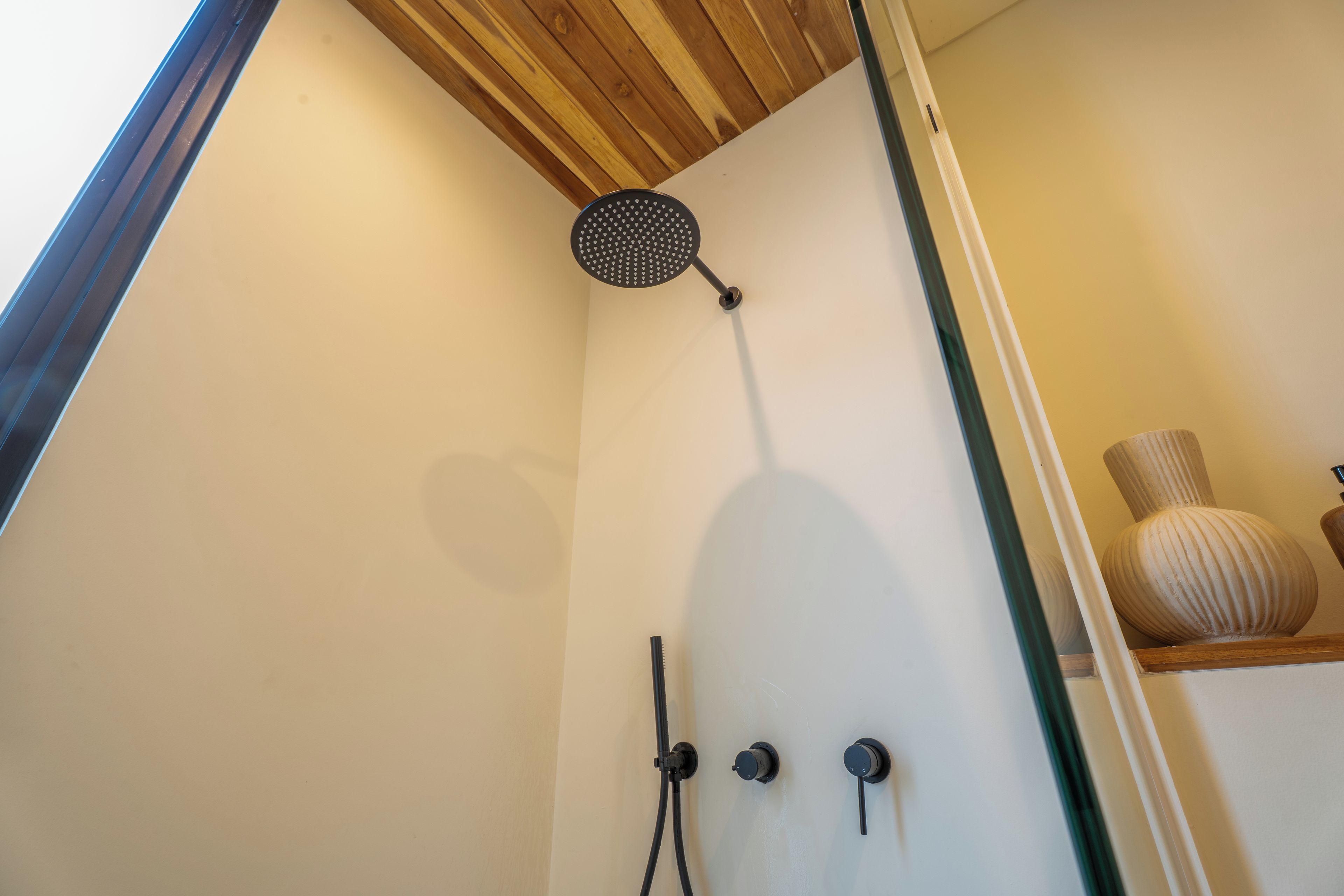 Een douche met een regendouchekop en twee knoppen, waarschijnlijk voor temperatuur en waterdruk, in een ruimte met een houten plafond en een witte wand.