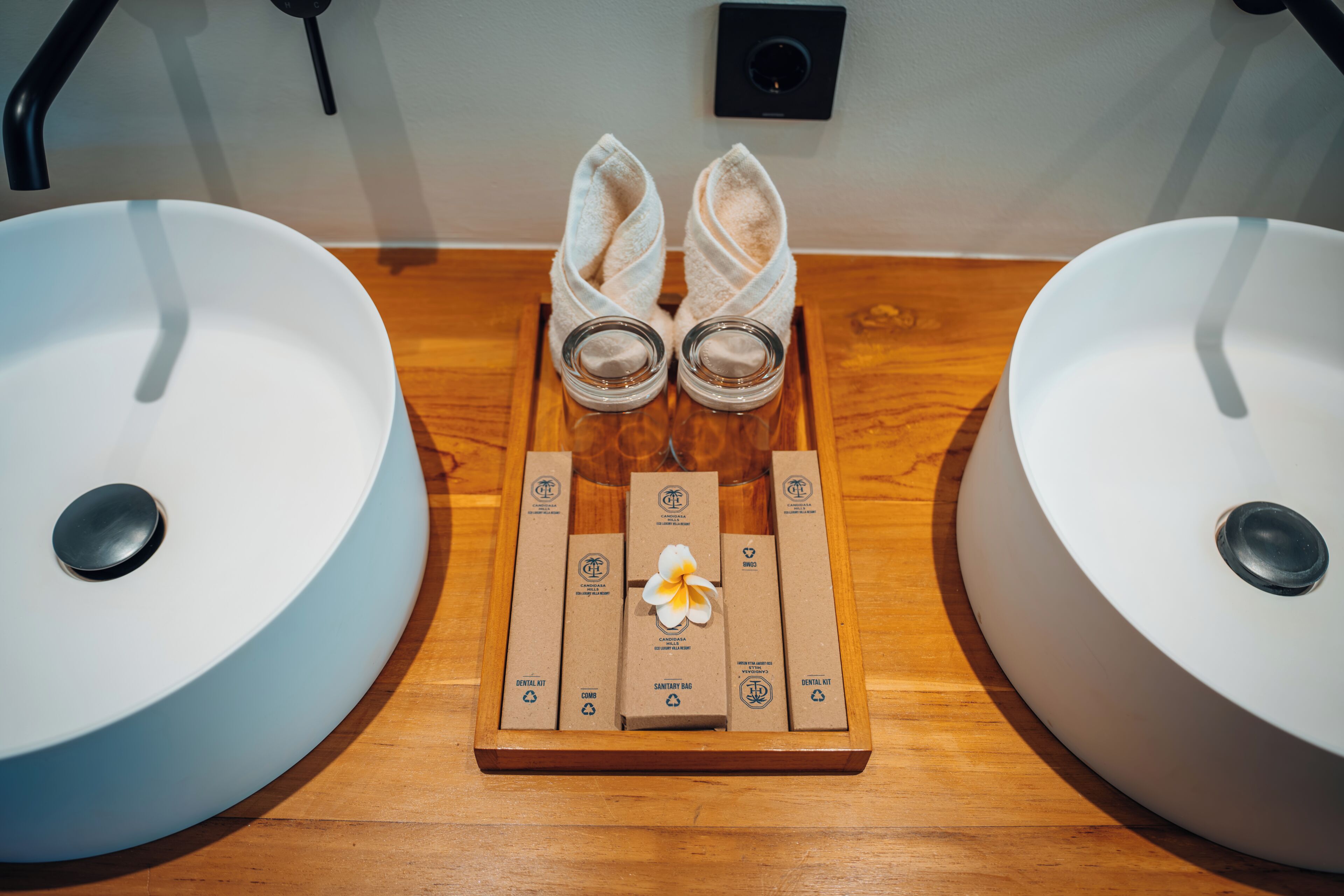 Un baño con dos lavamanos, una bandeja de madera con artículos de aseo y un par de pantuflas.