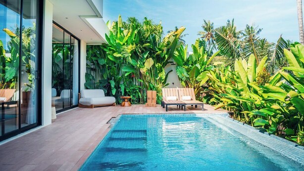 Un área de piscina moderna con sillones de descanso, una alberca y exuberantes plantas tropicales.