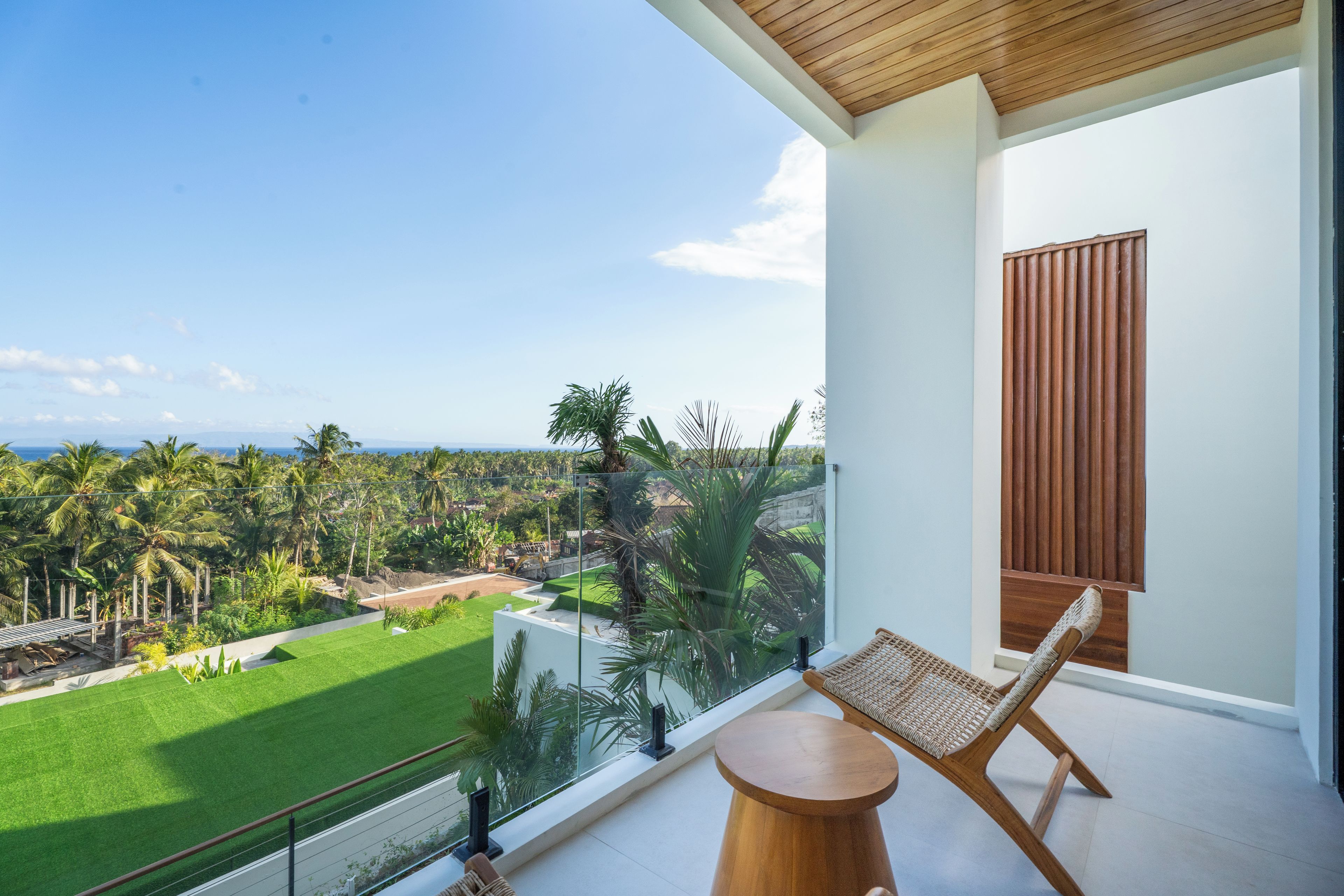 Un balcón con vista a un césped verde y palmeras, una silla de madera y una pequeña mesa redonda.