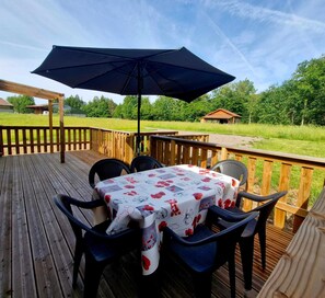 A wooden deck with a table and chairs, an umbrella, and a view of a grassy field and trees. - And why not?...rent a mobile home in the heart of nature (Ramonchamp)