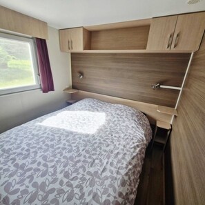 A bedroom with a bed, a window with curtains, and wooden cabinets. - And why not?...rent a mobile home in the heart of nature (Ramonchamp)