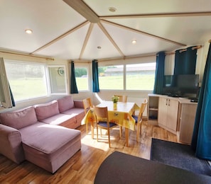 A cozy room with a sofa, a dining table with chairs, a TV, and a view of the outdoors. - And why not?...rent a mobile home in the heart of nature (Ramonchamp)